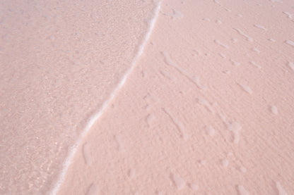 Fine art photography featuring a gentle wave lapping across a pink sand beach. The water is clear and small salty bubbles are visible across the sand, creating an organic design.